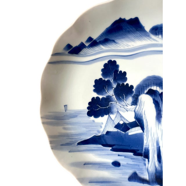 This hand-painted blue-and-white porcelain plate features a tranquil village landscape beneath rolling mountains, rendered...