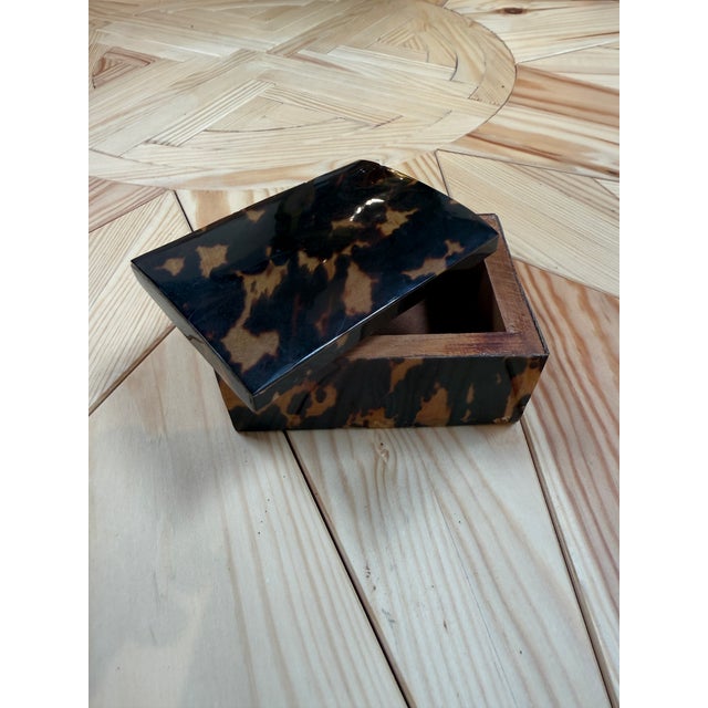 A compact rectangular box finished in a glossy faux-tortoiseshell pattern of mottled black, brown, and amber tones. The...