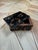 A compact rectangular box finished in a glossy faux-tortoiseshell pattern of mottled black, brown, and amber tones. The...