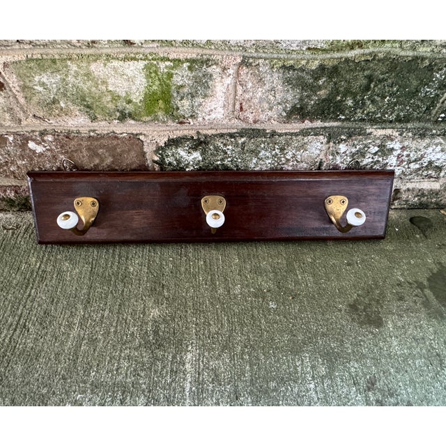 Wall hung trio of brass and white enamel hooks on piece of rich dark wood. This traditional wall hung coat rack has the...