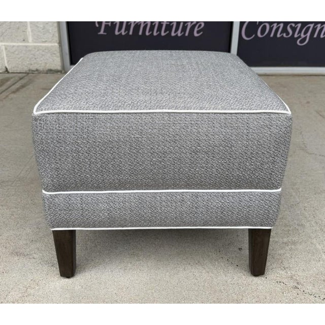 This Ethan Allen gray upholstered ottoman features a textured fabric, crisp white piping, and dark tapered wood legs for a...