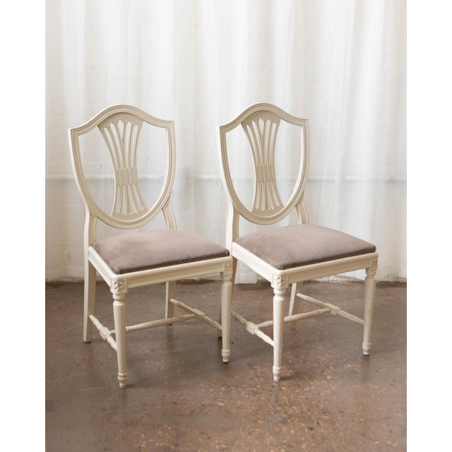 A well-composed set of seven Swedish Gustavian-style dining chairs in antique white painted frames with velour upholstered...