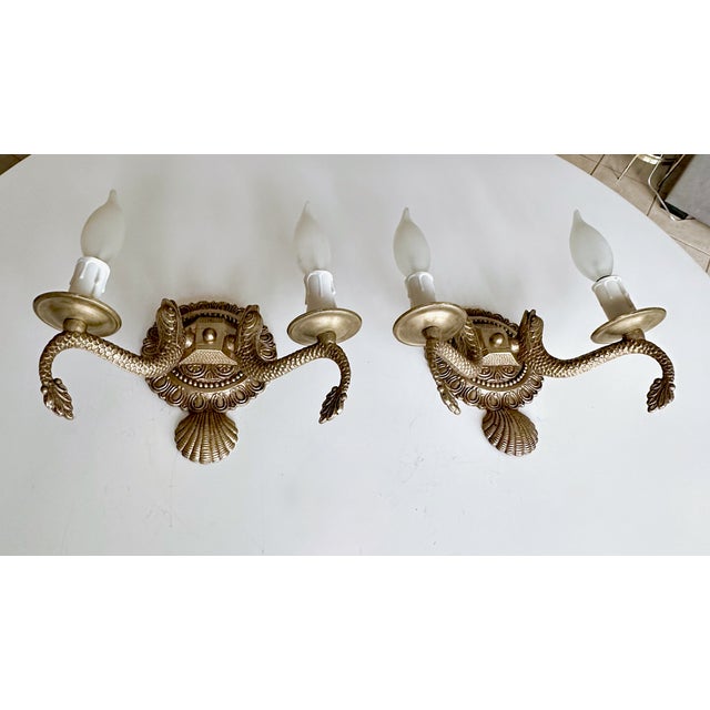 Pair of European two-light neoclassic style brass wall sconces with dolphin and claim shell motif. Expertly crafted with...