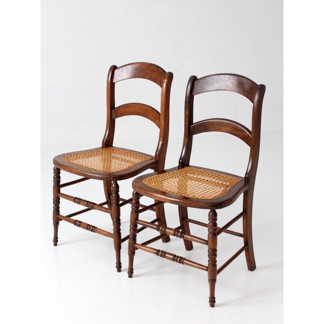 A handsome pair of early 20th-century ladderback dining chairs featuring hand-caned seats, turned front legs, and turned...