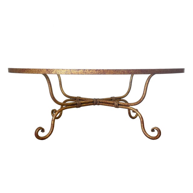 This is a beautiful Hollywood Regency styled oval coffee table featuring a hand-forged iron frame with a distressed gilt...