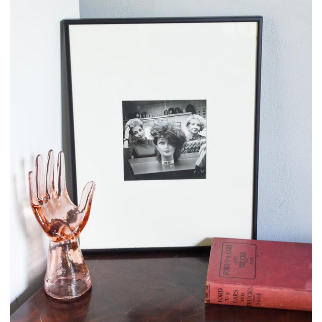 A 16 x 20 inch framed black-and-white photograph capturing a candid, warmly funny scene inside a Washington, DC wig shop:...