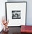 A 16 x 20 inch framed black-and-white photograph capturing a candid, warmly funny scene inside a Washington, DC wig shop:...