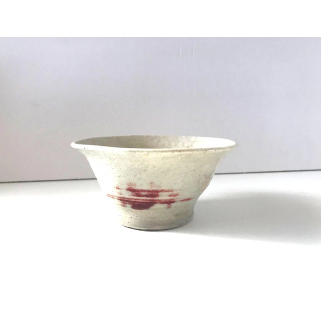 A vintage small hand-thrown ceramic bowl in a white or light grey glaze with a deep red detail on one side and crazing...