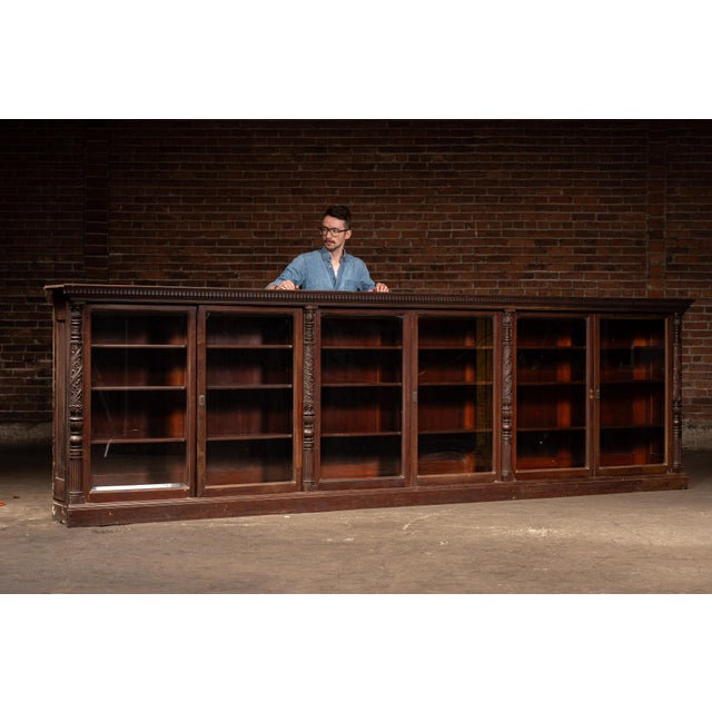 Expansive multi-bay library cabinet featuring a continuous run of glass-front display sections framed by richly carved...