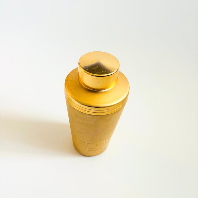 A Mid Century gold-tone aluminum cocktail shaker. The shaker is matte gold in coloring with shiny gold bands and a cap for...