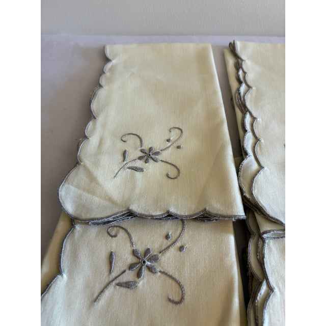 This set of 8 ivory napkins features delicate gray floral embroidery in one corner and a matching scalloped edge finished...