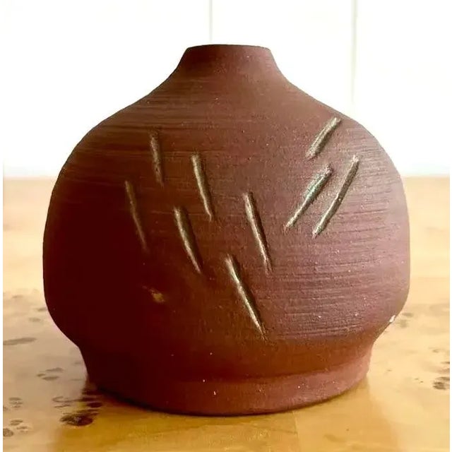 This is a small, hand-thrown ceramic bud vase with a rounded form and narrow neck. Its matte reddish-brown clay surface is...