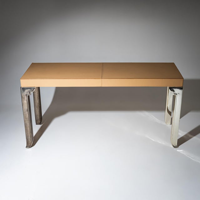 Long rectangular console table on straight stainless steel legs with decorative profiling. The top is covered in beige...