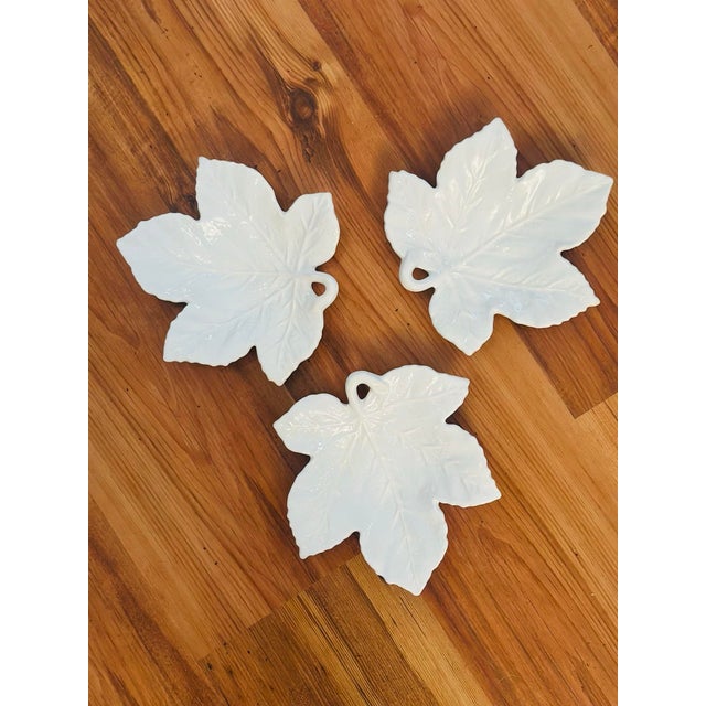 These are white porcelain leaf-shaped dishes with a glossy finish. The leaves have detailed veining and a small loop-like...