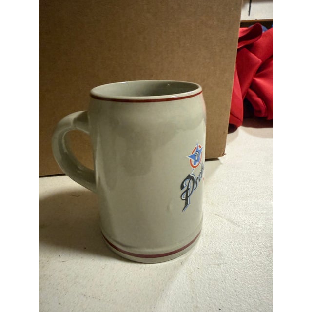 Set of two mugs. 1. Hofbräuhaus (HB) 1-Liter Lidded Stein: Features the classic blue crowned HB logo on a traditional gray...