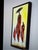 In this dark framed folk art lithograph painting depicting stylized Maasai figures of women in vibrant red attire in a...