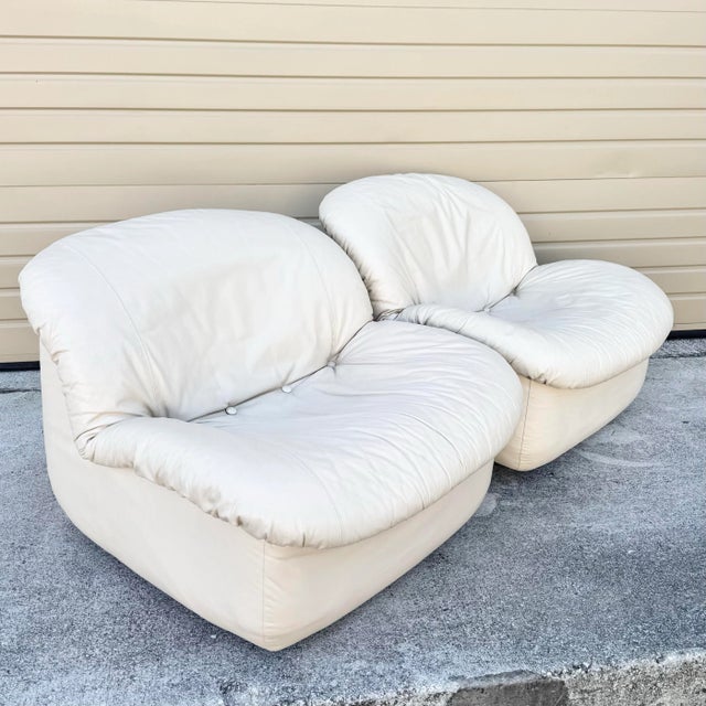 A sleek pair of Postmodern swivel lounge chairs in soft cream leather, featuring plush tufted cushions and sculpted low-...