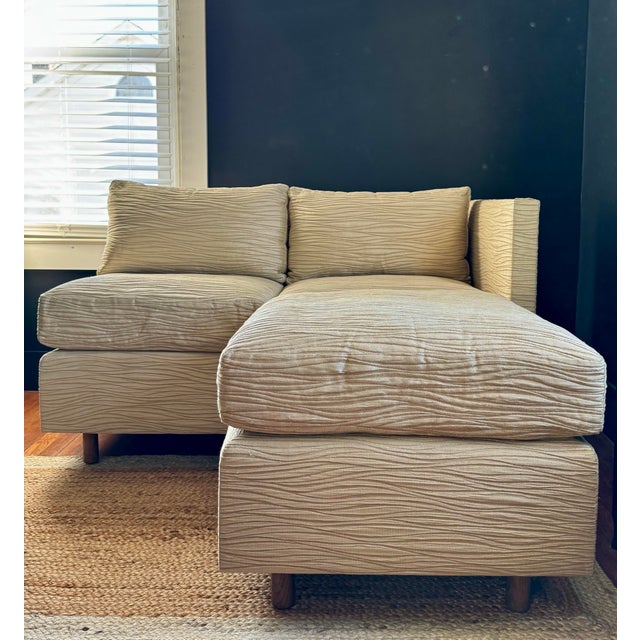 Small scale even arm occasional sofa with ottoman in textured, champagne colored upholstery on small cylindrical wooden...