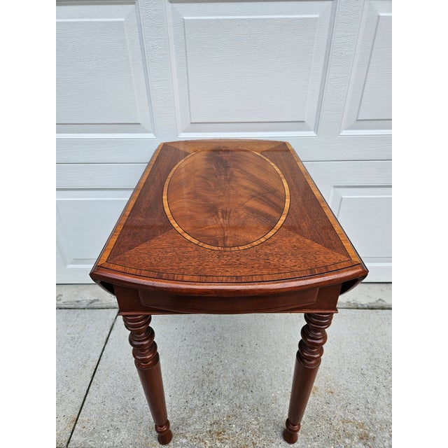 This beautiful one drawer Pembroke drop-leaf side table is by Baker Furniture and features an oval crotch mahogany veneer...