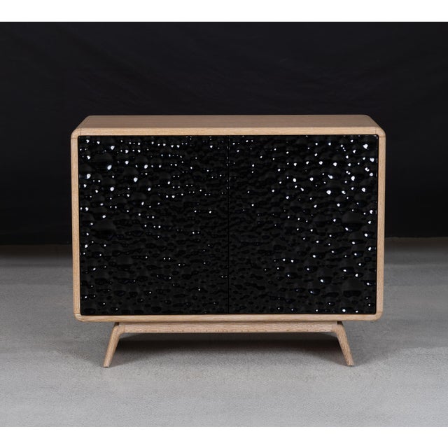 A modern accent cabinet pairs a low, rounded case with a broad, splayed base. Deeply patterned black cast-glass doors...