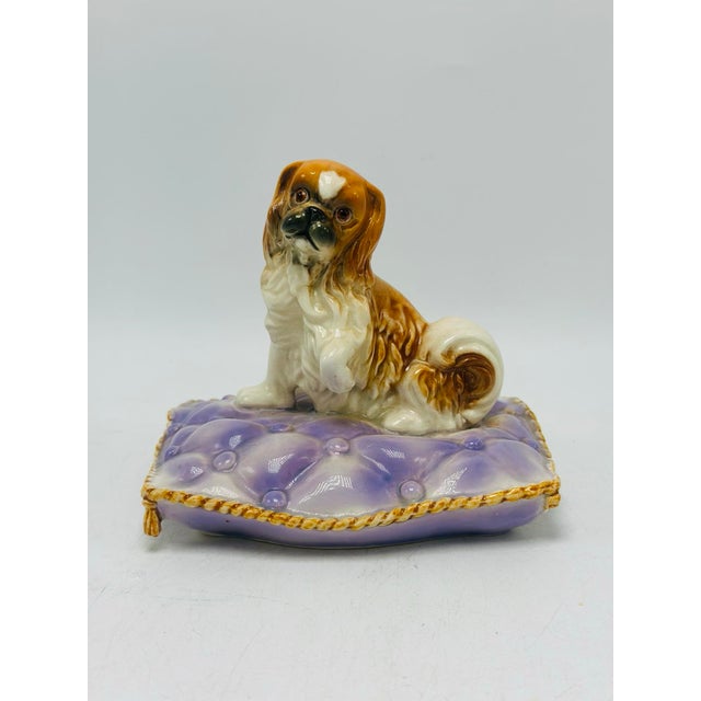 This is a vintage ceramic dog figurine featuring a small spaniel resting on a decorative tufted lavender cushion with gold...
