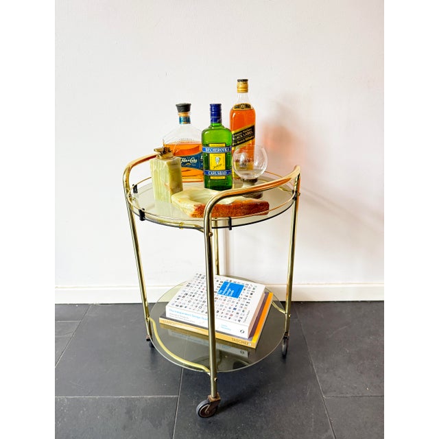 A vintage bar cart in a mid-century modern style, featuring a brass frame, two removable smoked glass shelves and four...