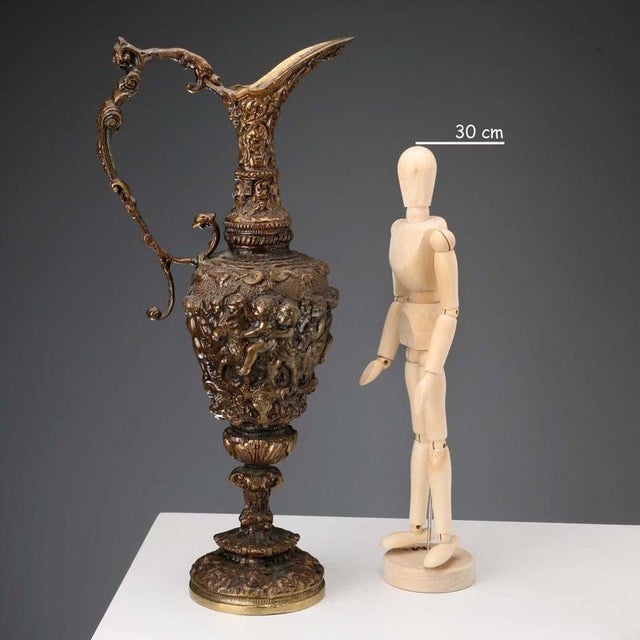 A bronze jug carved in bas-relief. The belly features scenes with cherubs. The jug features a handle with a mythological...