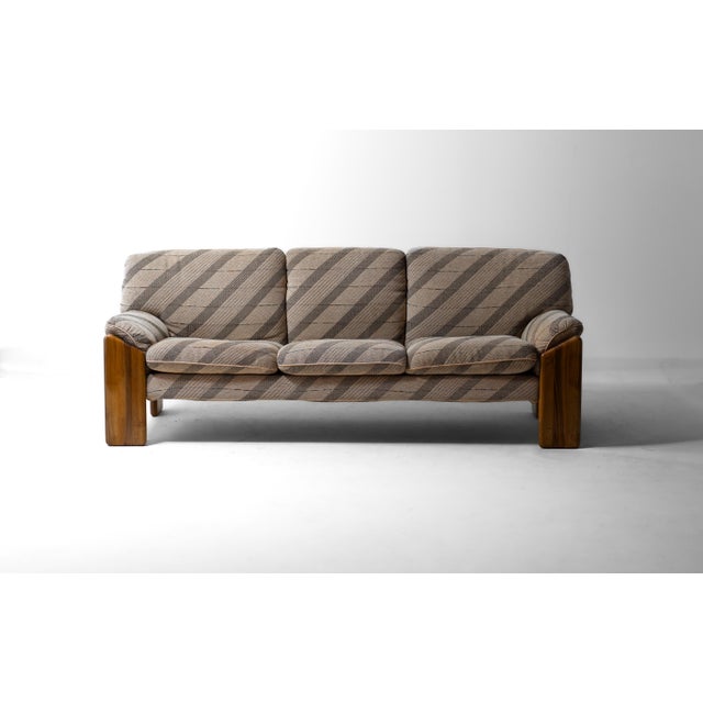 Three-seater sofa with a solid walnut frame. Four broad, rounded legs carry a curved back rail that stays visible behind...
