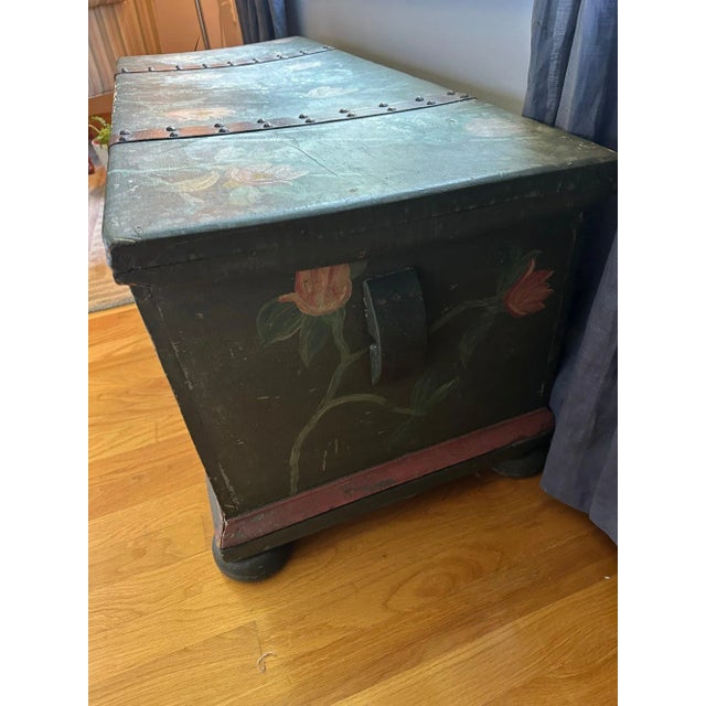 A rustic antique wooden storage chest with charming hand-painted decoration. The piece features a deep green base accented...