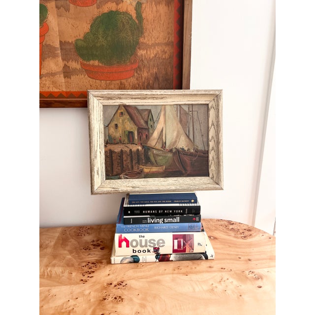 charming mid-century oil painting depicting sailboats moored in a picturesque harbor, with waterfront cottages and small...