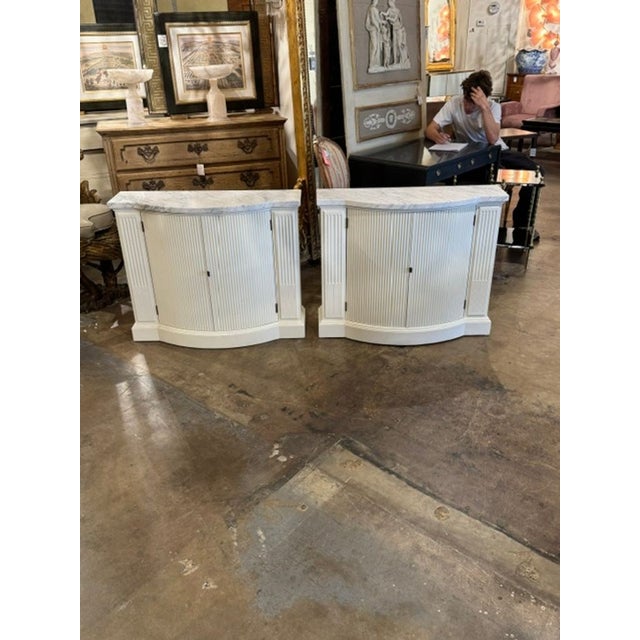 The product is a pair of vintage Italian white painted side cabinets with Carrara marble accents. The cabinets are...