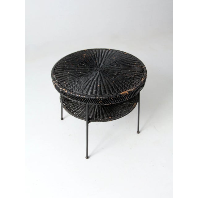 A mid-century black wicker side table with a two-tier design featuring round woven surfaces and slim metal legs. the airy...