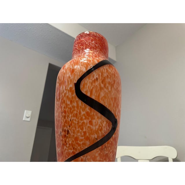 This hand-blown art glass vase features a vibrant orange body accented by a dramatic black applied swirl, creating a...