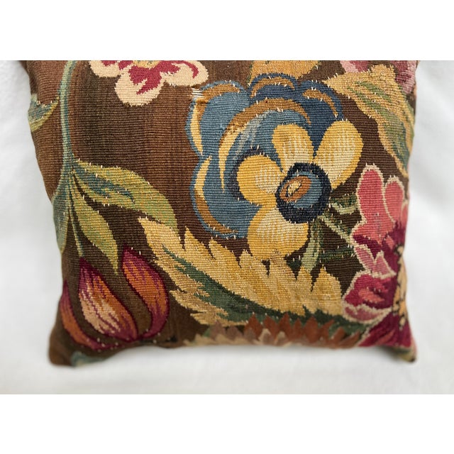 An exquisite antique-style pillow upholstered in richly woven 19th-century tapestry, set against a deep warm brown ground...