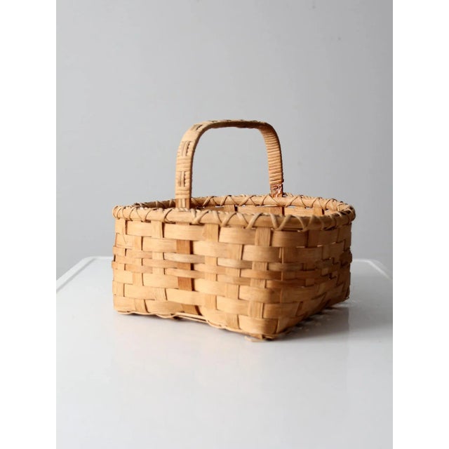 A vintage woven harvest basket featuring traditional splint weaving, a sturdy wooden top frame and handle, and an open-...