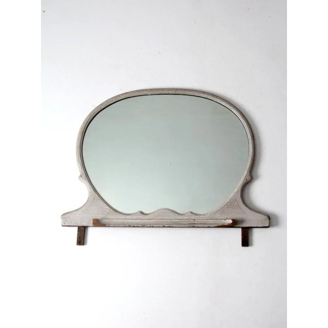 An antique oval mirror featuring a decorative wooden frame with a lipped base and original crackled white paint. The aged...