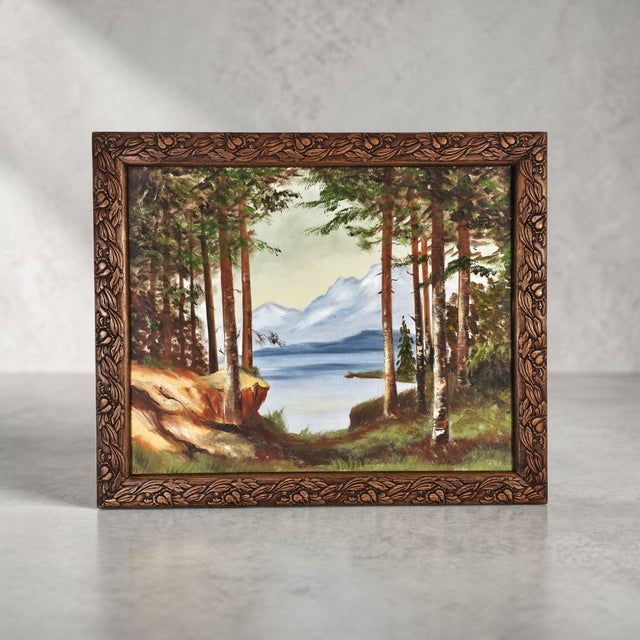 A vintage landscape oil painting depicting a mountain lake framed by tall pines, presented within a carved wood frame of...