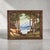 A vintage landscape oil painting depicting a mountain lake framed by tall pines, presented within a carved wood frame of...