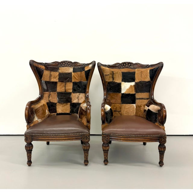 A distinctive pair of carved wood wingback armchairs upholstered in patchwork cowhide in rich shades of brown, black, tan,...