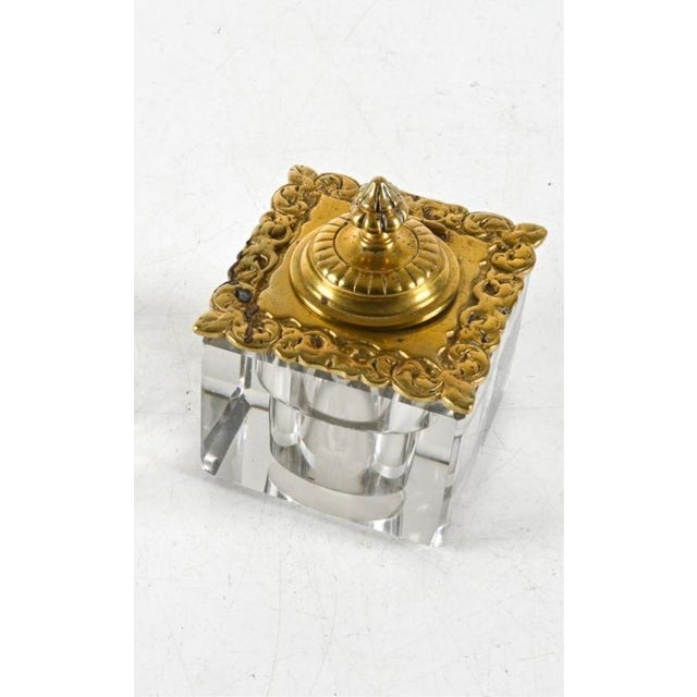 A square-cut crystal inkwell with thick, faceted glass walls, cylindrical well, gilt brass hinged lid with a pierced,...