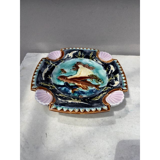 Square Majolica mermaid plate with shells on the corners, the mermaid sit on a flying fish, the border is decorated with...