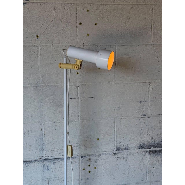 This minimalist floor lamp features an adjustable tubular shade mounted on a slim white metal standard. The sleek...