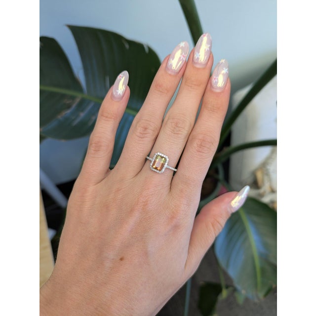 A bi-color tourmaline glows in shades of green and warm orange at the center of this emerald-cut ring, framed by a halo of...