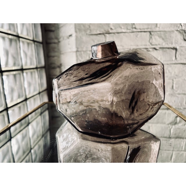 A striking, faceted hand-blown glass vase with a smoky amethyst hue. Its geometric, almost rock-like shape gives it a...