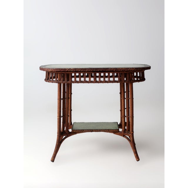 This early 20th-century American wicker accent table features an elongated oval top and a lower display shelf, each fitted...