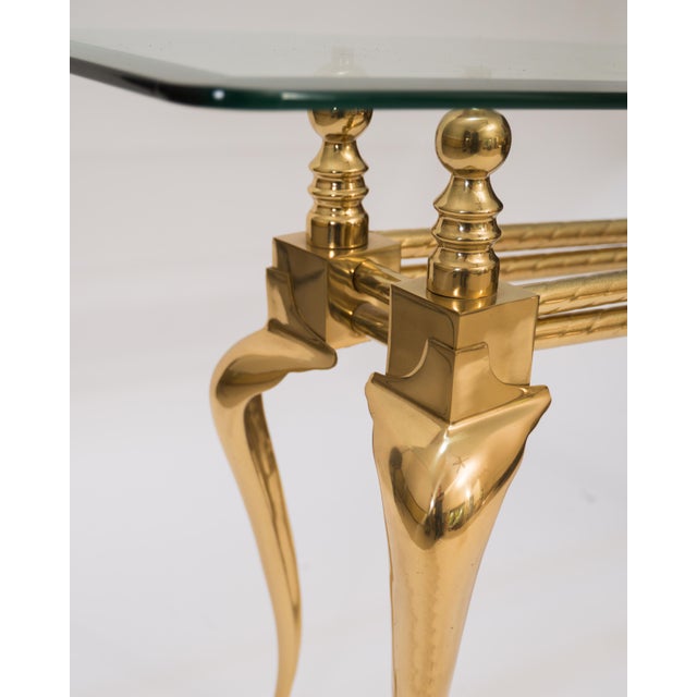 This 1970s console table features a clear thick rectangular glass top with polished edges supported by an ornate brass...