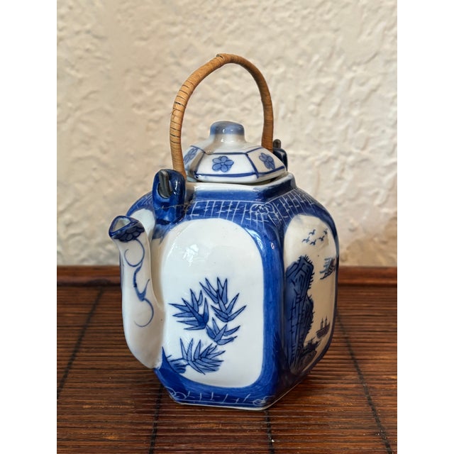 A blue and white Japanese landscape tea pot hand decorated with trees and boats. Handle is made of rattan wrapped metal....