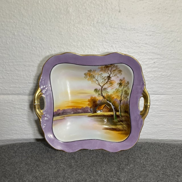 Hand painted landscape scene of a lone cottage with lake and swan. Purple border with dual handles trimmed in gold....