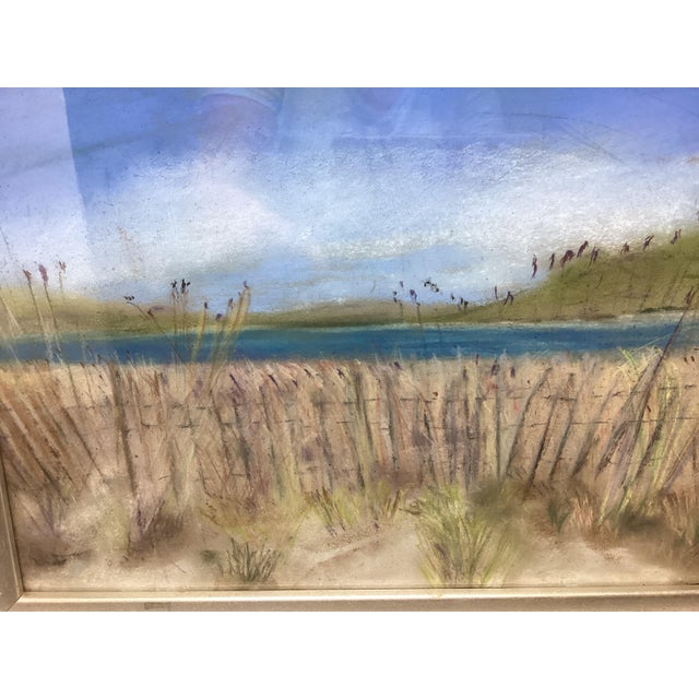 This framed seascape pastel painting features sand dunes in the foreground and a calm blue ocean under a clear sky. This...