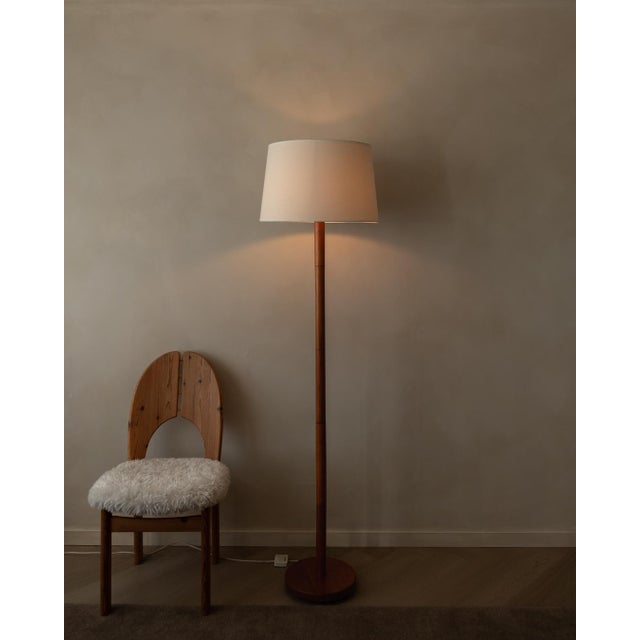 A calm, elegant vintage floor lamp with a slender teak-toned wooden stem, round base, and a clean fabric shade that gives...